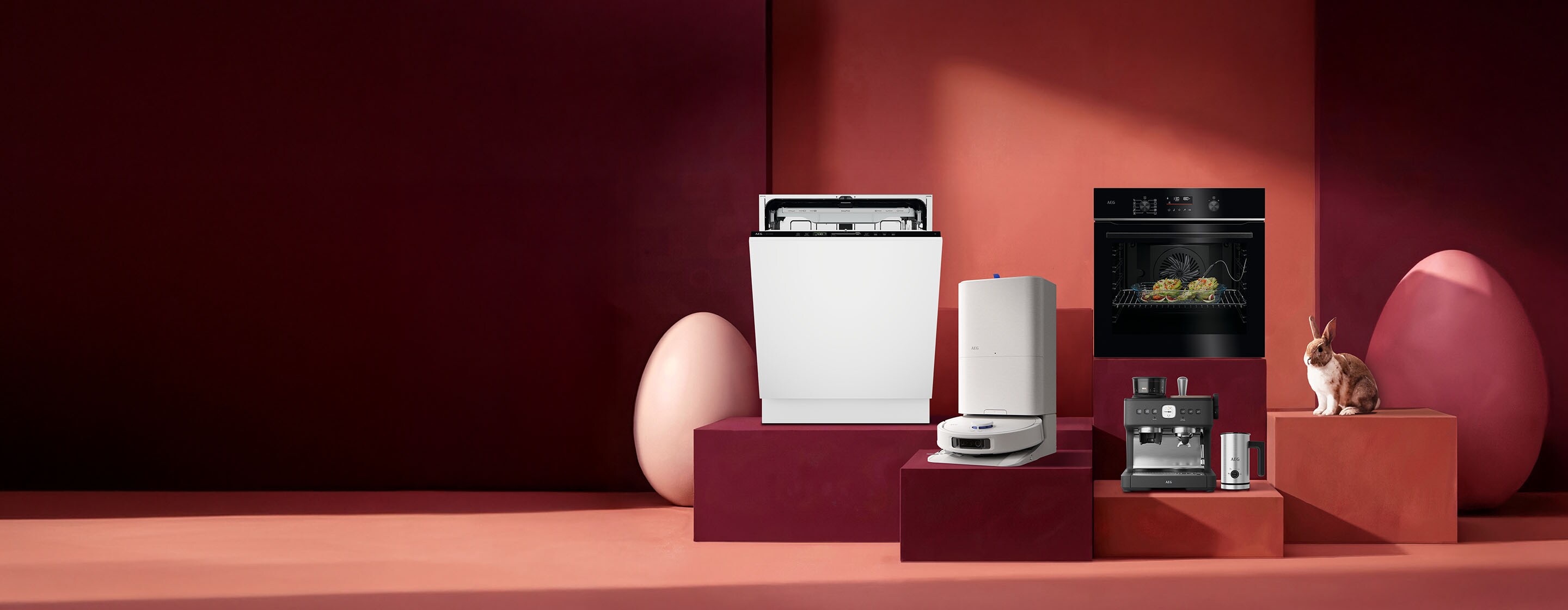 Mehrere Haushaltsgeräte, darunter ein Geschirrspüler, ein Saugroboter, ein Luftreiniger, ein Backofen und eine Kaffeemaschine, stehen dekorativ auf farbigen Podesten in einer rot‑rosafarbenen Studio­umgebung; rechts sitzt ein kleines Kaninchen