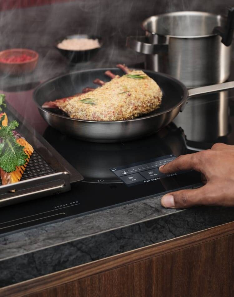 Ein Finger berührt das Bedienfeld eines Induktionskochfelds, während Essen zubereitet wird.