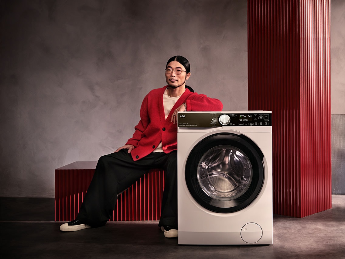 Mann in rotem Cardigan lehnt an AEG Waschmaschine der 9000er Serie vor grauem Hintergrund