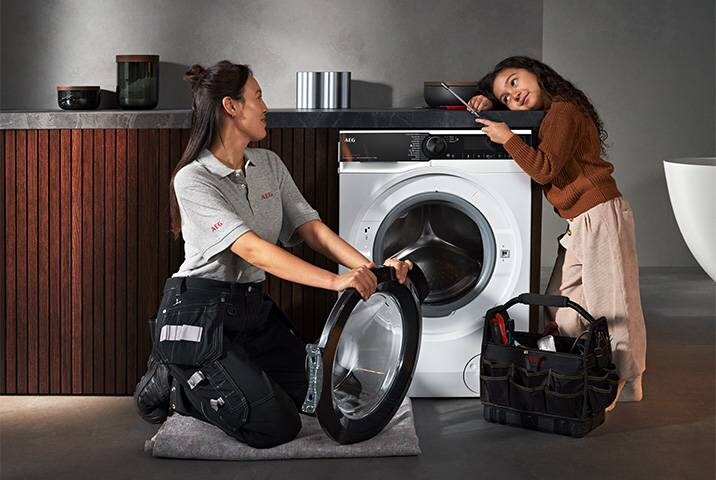 AEG Servicetechnikerin tauscht Türdichtung an AEG Waschmaschine aus, Kundin schaut interessiert zu